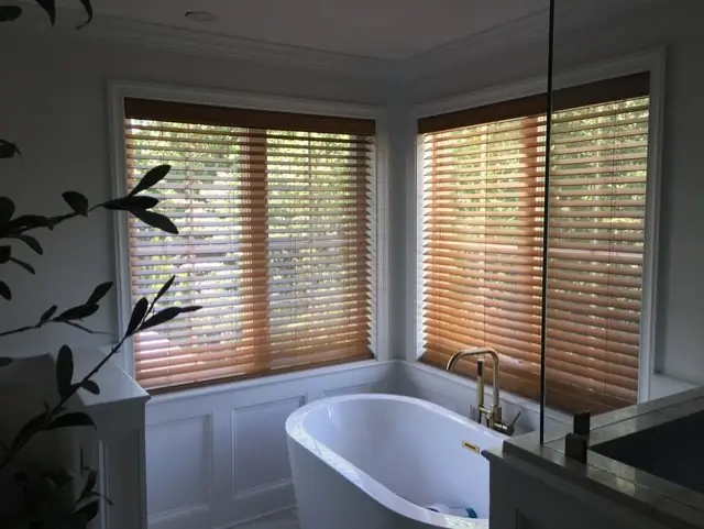 Honey-toned wood blinds in a bathroom corner beside a freestanding tub